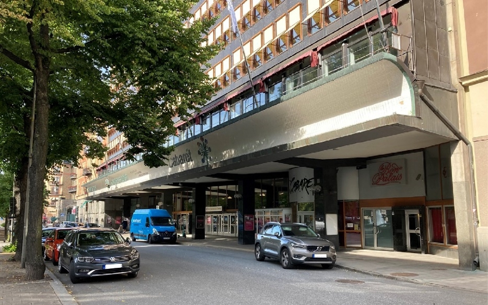 Ett flervåningshus där entrén har ett skärmtak. Bilar står parkerade på gatan vid entrén.